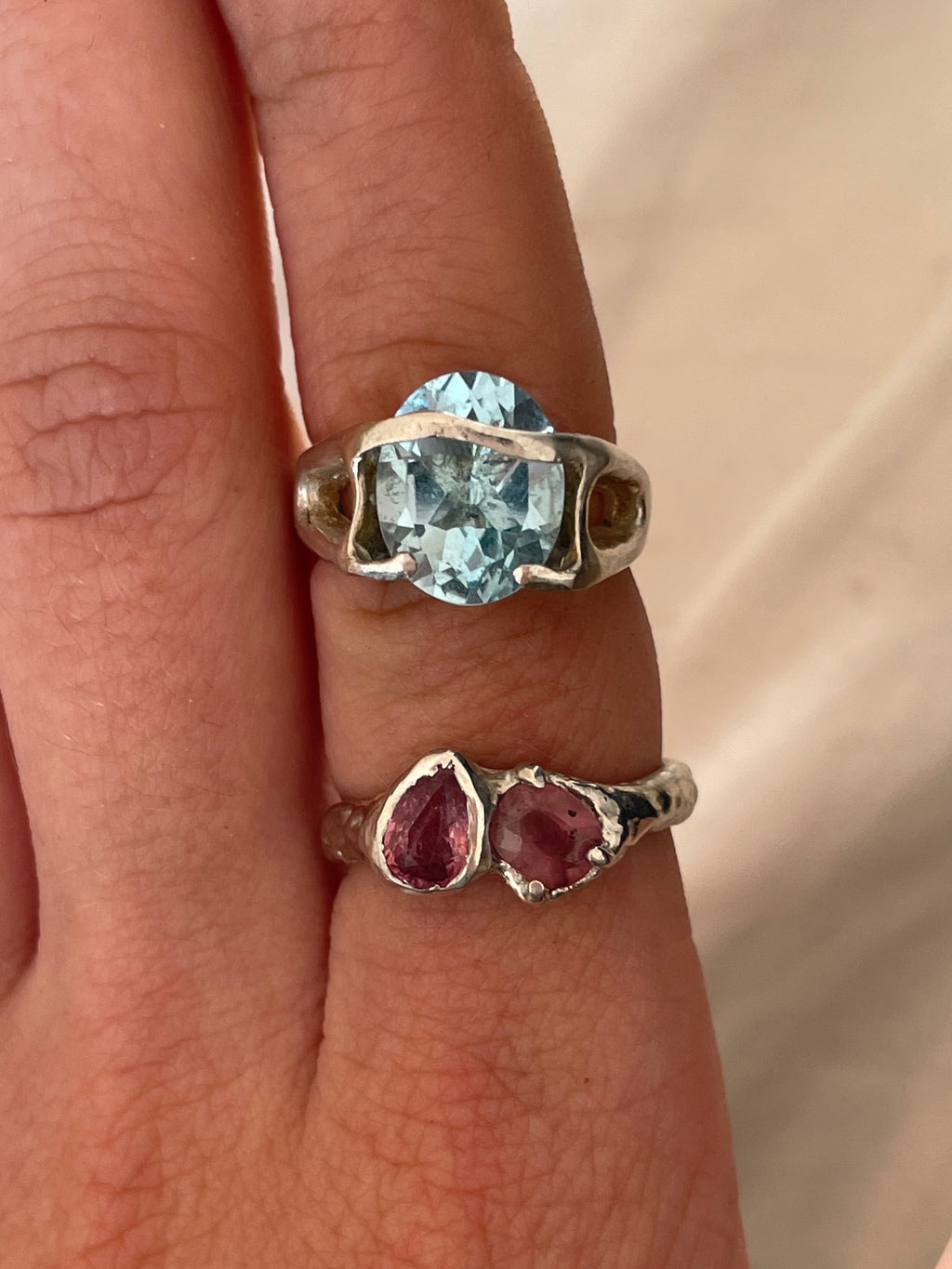 Two silver rings with gemstones on a person's finger . One ring is silver ring with two pink sapphires and other is with blue topaz.